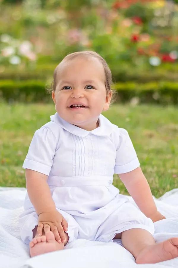Barboteuse Baptême bébé garçon Augustin en piqué de coton blanc et dentelle de coton. Création française Fil de Légende. Photo d'un bébé assis portant la barboteuse.