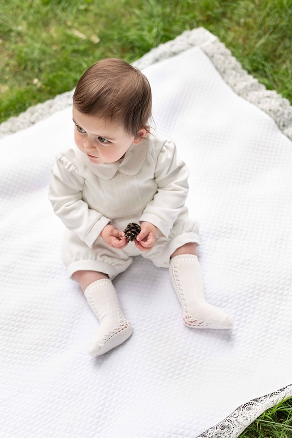 Barboteuse baptême d'hiver bébé garçon Baptiste en velours blanc cassé et dentelle de Calais. Doublure polaire. Création française Fil de Légende. Photo de la barboteuse portée par un bébé garçon assis.