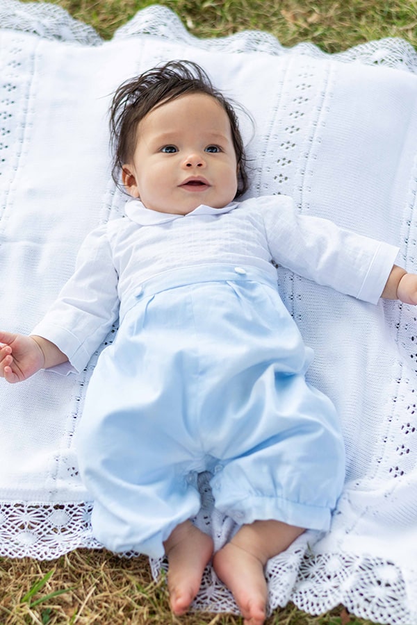 Barboteuse cérémonie bébé garçon Hugo. Corsage en coton brodé blanc. Pantalon en coton bleu ciel. Création française Fil de Légende. Photo de la barboteuse portée par un bébé garçon allongé.