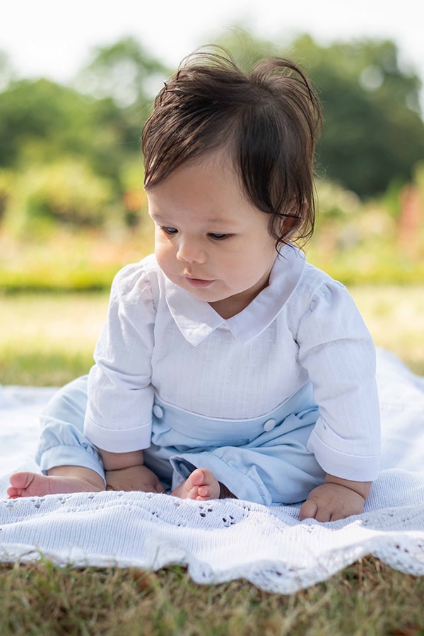 Barboteuse cérémonie bébé garçon Hugo. Corsage en coton brodé blanc. Pantalon en coton bleu ciel. Création française Fil de Légende. Photo de la barboteuse portée par un bébé garçon brun.