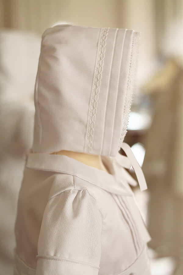 Bonnet de baptême bébé garçon Augustin en piqué de coton blanc et dentelle de coton. Création française Fil de Légende. Photo du bonnet porté avec la barboteuse assortie.