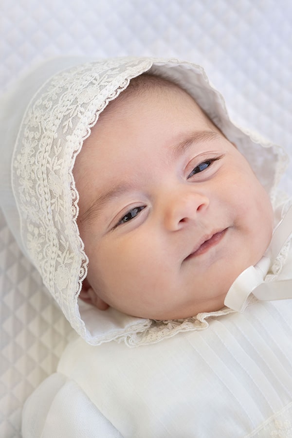 Bonnet de baptême bébé fille Gloria en mousseline et dentelle blanc cassé. Création française Fil de Légende. Photo du bonnet porté par un bébé fille.
