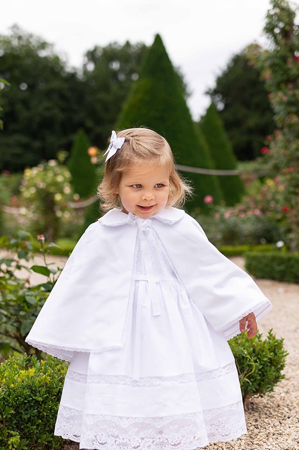 Cape de baptême bébé fille en coton blanc et galon de dentelle. Création française Fil de Légende. Photo de la cape portée par-dessus une robe de baptême.