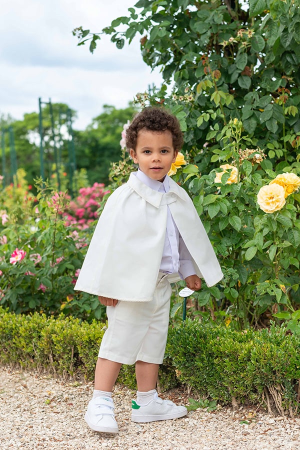 Cape de baptême bébé garçon Oscar en coton blanc cassé et galons de dentelle de coton. Création française Fil de Légende. Photo de la cape portée par un garçon dans un jardin.