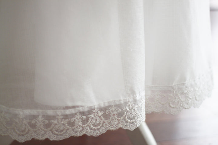 Cape de baptême longue traditionnelle pour baptême en mousseline blanc cassé et dentelle fine. Se ferme avec un large ruban. Fait main en France. Gros plan sur le bas de la cape.