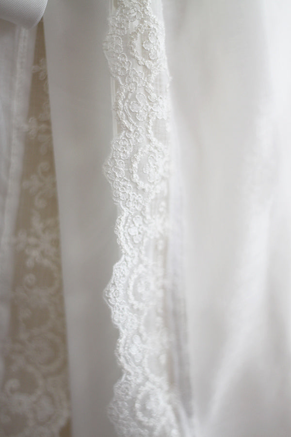 Cape de baptême longue traditionnelle pour baptême en mousseline blanc cassé et dentelle fine. Se ferme avec un large ruban. Fait main en France. Gros plan sur bordure.