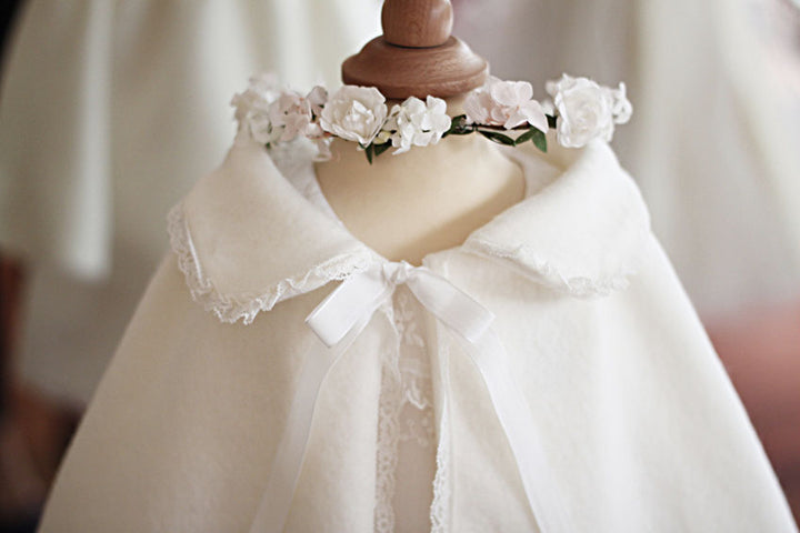 Cape d'hiver bébé fille pour cérémonie  ou baptême en lainage blanc cassé et bordure dentelle. Photo avec couronne de fleurs blanches.