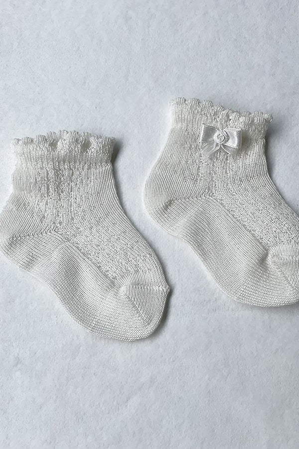 Chaussettes bébé fille en coton blanc cassé ajourées avec un bord dentelé et un petit noeud en satin sur le côté. Parfaites pour être portées avec une robe de cérémonie bébé ou une robe de baptême.