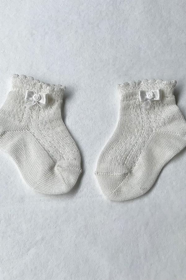 Chaussettes bébé fille en coton blanc cassé ajourées avec un bord dentelé et un petit noeud en satin sur le côté. Parfaites pour être portées avec une robe de cérémonie bébé ou une robe de baptême.