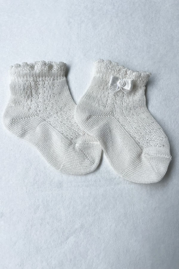 Chaussettes bébé fille en coton blanc cassé ajourées avec un bord dentelé et un petit noeud en satin sur le côté. Parfaites pour être portées avec une robe de cérémonie bébé ou une robe de baptême.