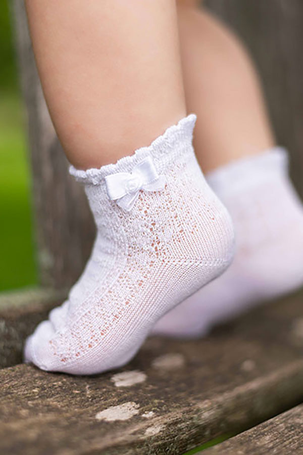 Chaussettes bébé fille en coton blanc ajourées avec un bord dentelé et un nœud en satin blanc sur le côté. A porter avec une robe de cérémonie bébé ou une robe de baptême - vue sur coté gauche
