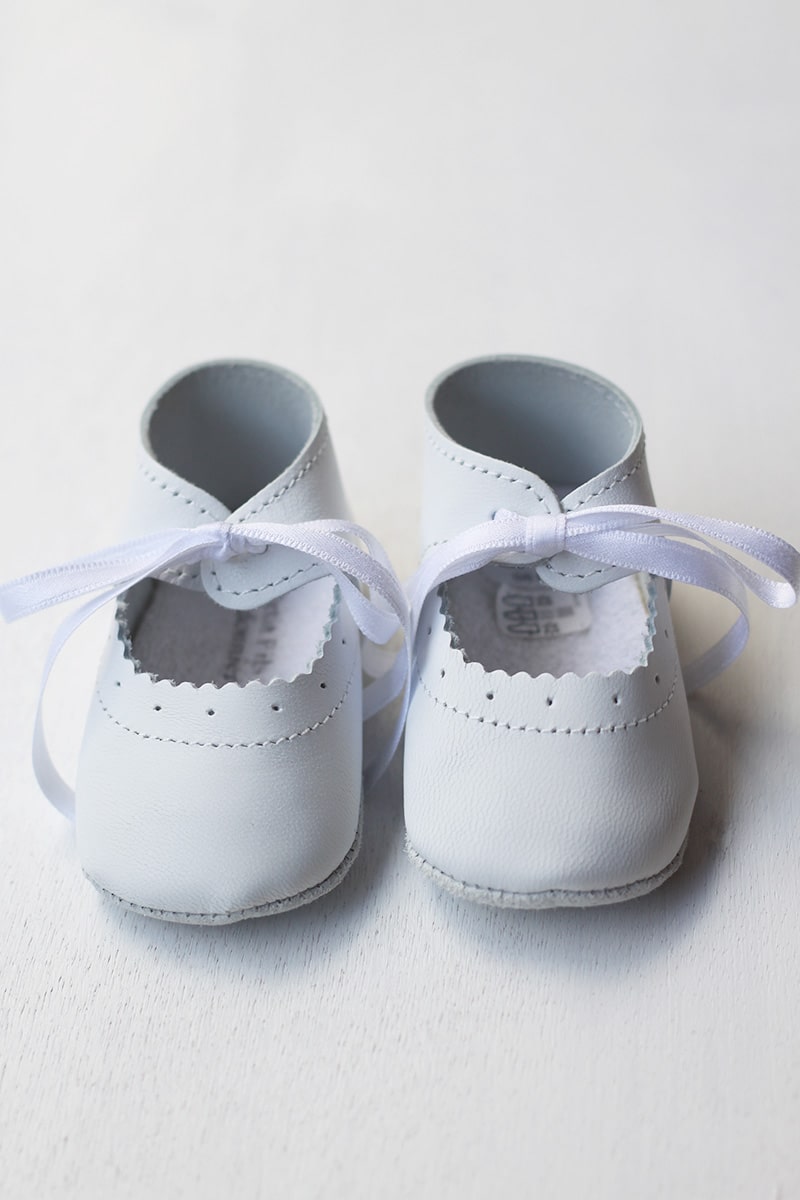 Chaussons ballerines bébé fille en cuir blanc pour cérémonie ou baptême. Se ferme en nouant un ruban de satin. Magasin Fil de Légende Paris, Neuilly-sur-Seine.