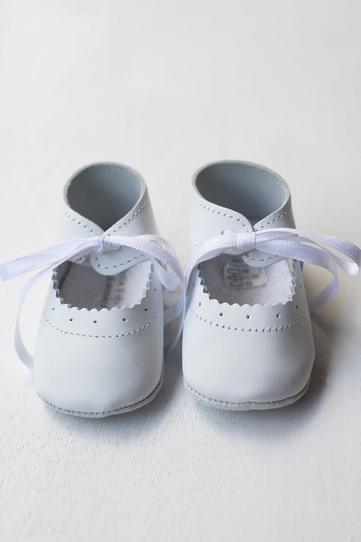 Chaussons ballerines bébé fille en cuir blanc pour cérémonie ou baptême. Se ferme en nouant un ruban de satin. Magasin Fil de Légende Paris, Neuilly-sur-Seine.