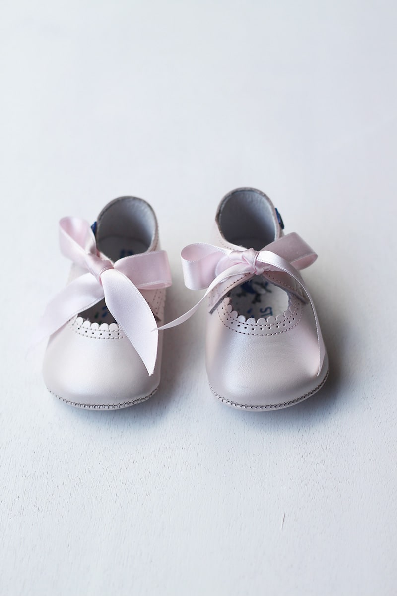 Ballerines bébé fille en cuir rose pâle avec une bordure dentelée pour cérémonie ou fête. Les chaussons se ferment avec un noeud en satin rose à nouer. Vue éloignée. 