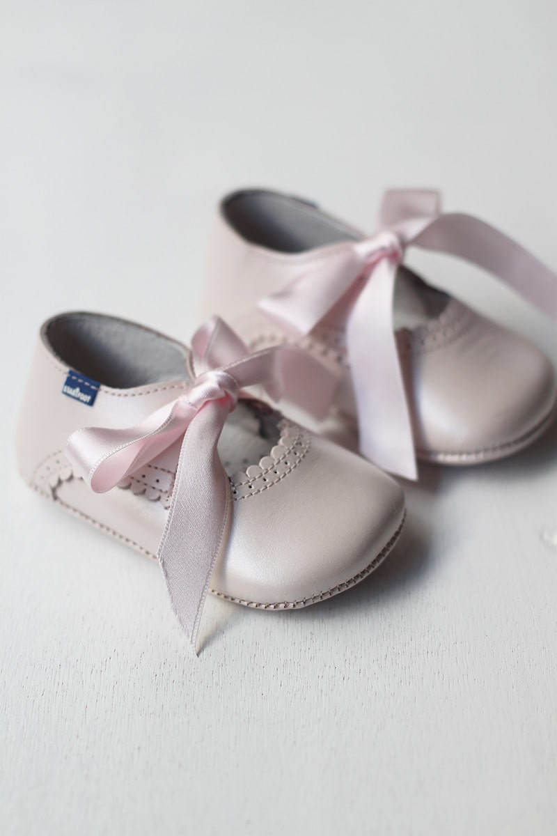 Ballerines bébé fille en cuir rose pâle avec une bordure dentelée pour cérémonie ou fête. Les chaussons se ferment avec un noeud en satin rose à nouer. Vue de côté. 