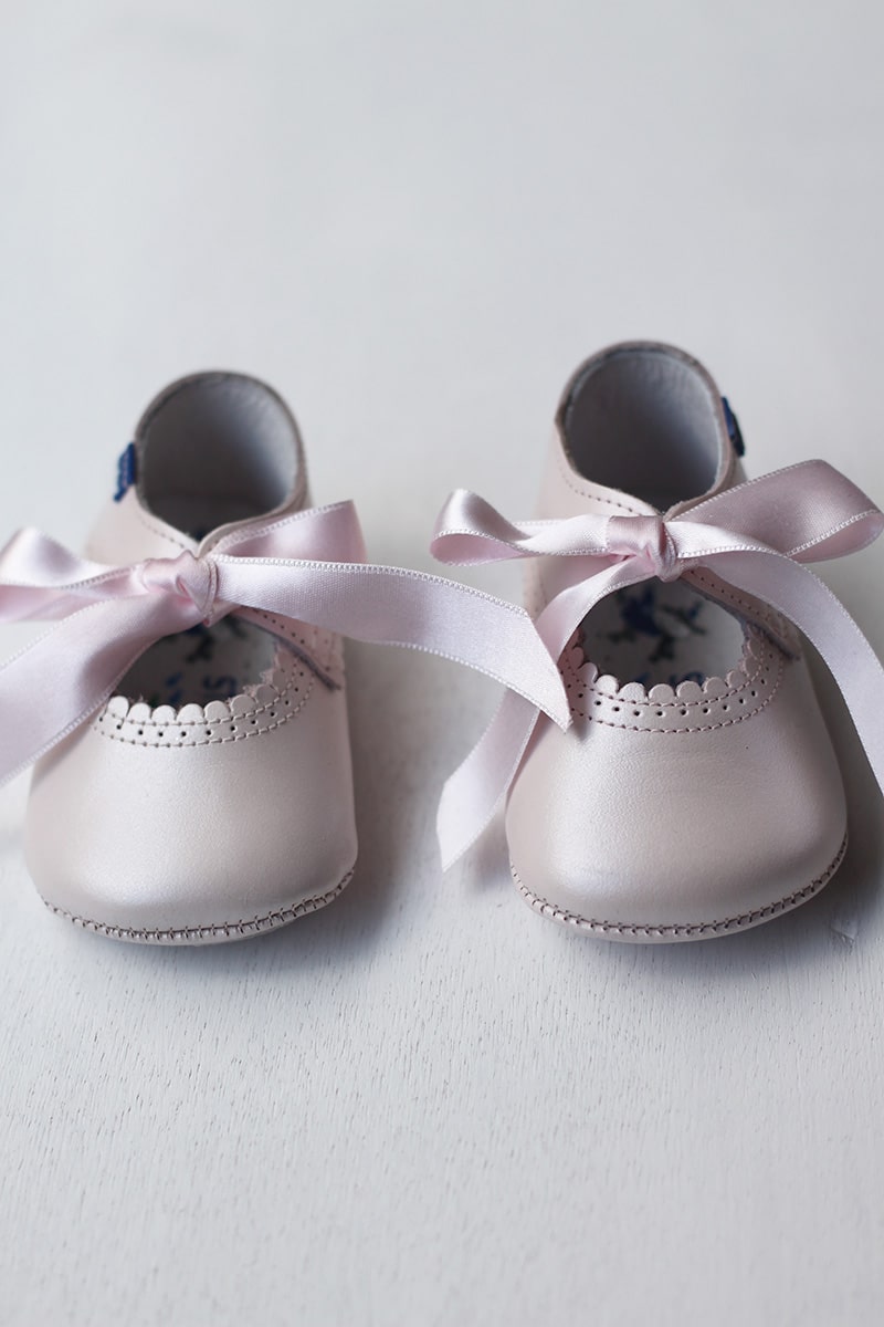 Ballerines bébé fille en cuir rose pâle avec une bordure dentelée pour cérémonie ou fête. Les chaussons se ferment avec un noeud en satin rose à nouer. Magasin Fil de Légende Paris, Neuilly-sur-Seine.