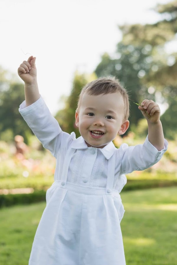 Ensemble baptême bébé garçon Alexandre avec chemise et salopette en lin blanc et dentelle de coton. Création française Fil de Légende. Photo de l'ensemble porté par un petit garçon qui joue.