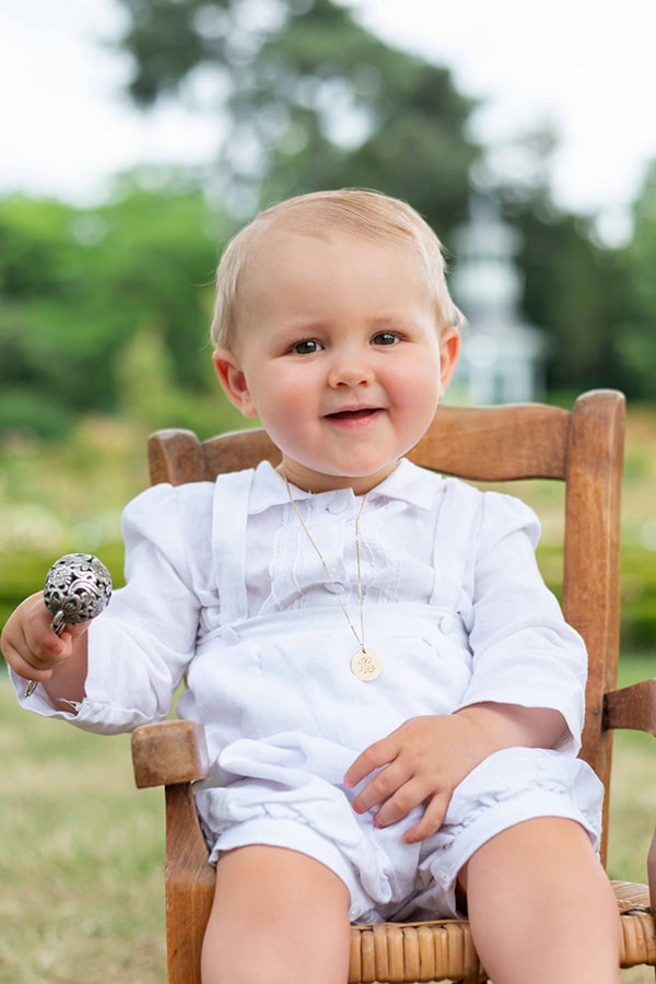 Ensemble baptême bébé garçon Alexandre avec chemise et salopette en lin blanc et dentelle de coton. Création française Fil de Légende. Photo de l'ensemble porté par un bébé garçon avec un hochet.