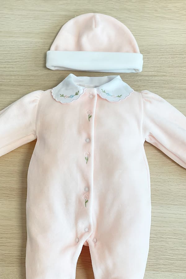 Ensemble de naissance bébé fille Eva en velours de coton rose pâle, avec pyjama à col blanc brodé de fleurs et bonnet assorti à revers blanc, présenté sur fond bois clair – Fil de Légende.