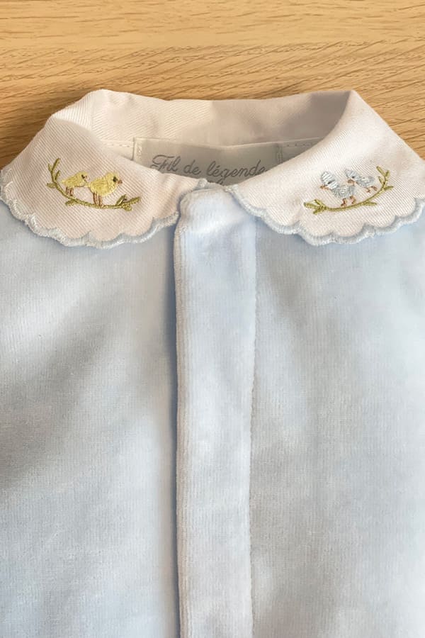 Gros plan sur le col blanc festonné du pyjama bébé garçon Elio en velours de coton bleu ciel, délicatement brodé de petits oiseaux jaunes et bleus posés sur des branches – Fil de Légende.
