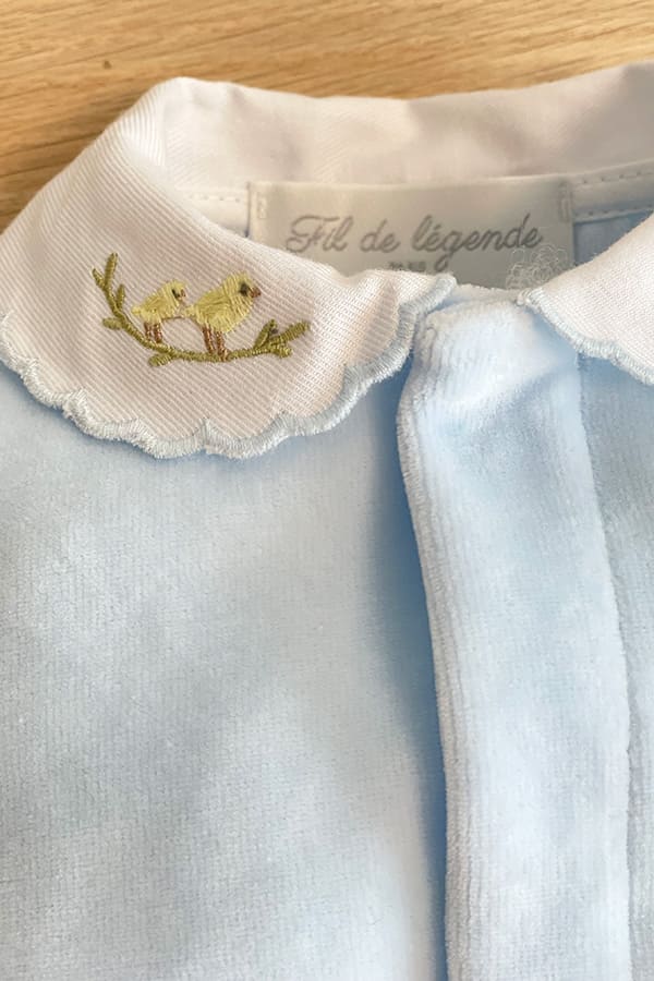 Gros plan sur la broderie de deux petits oiseaux jaunes posés sur une branche, ornant le col blanc festonné du pyjama bébé garçon Elio en velours de coton bleu ciel – Fil de Légende.