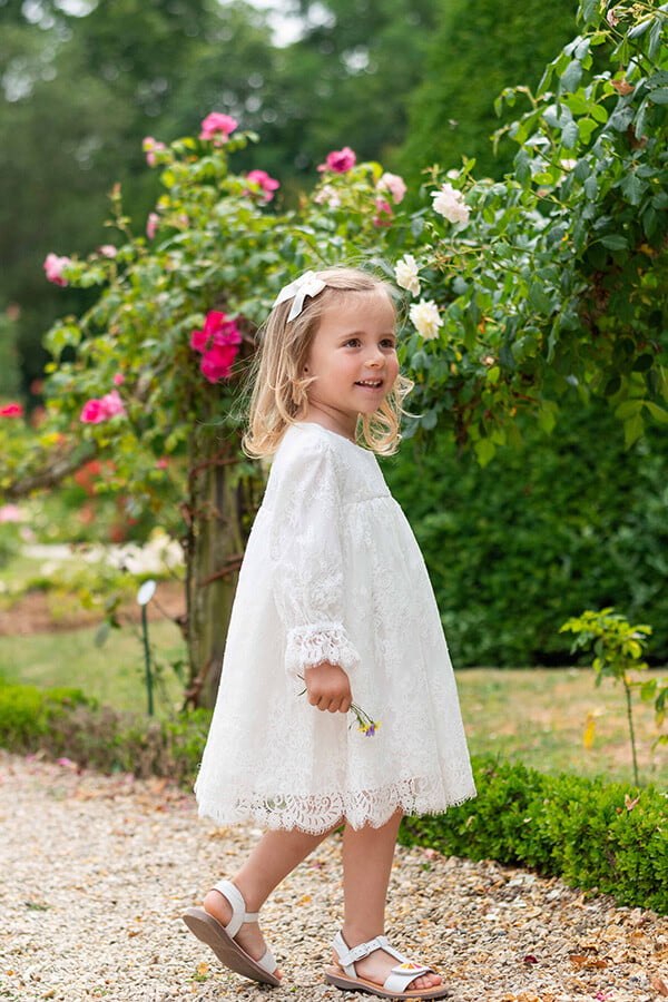 Petite fille portant une robe de baptême ou de cérémonie en dentelle italienne, doublée de coton, modèle printemps à manches longues. Création Fil de Légende, tenue de cérémonie pour bébé et petite fille.
