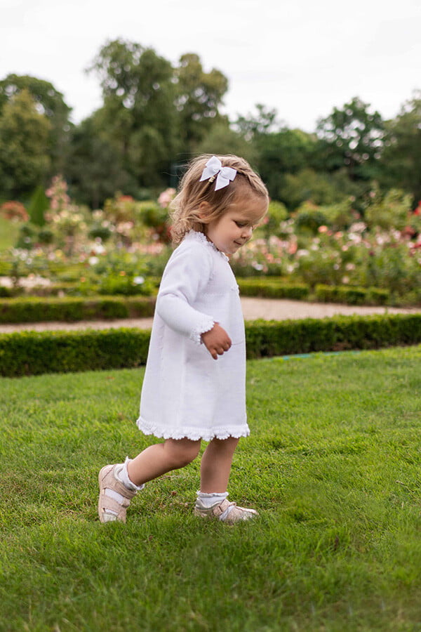 Robe baptême fille blanc cassé en maille tricotée avec bordures faites au crochet à la main. Robe en lainage. Création française Fil de Légende. Photo de la robe portée par une petite fille qui marche.