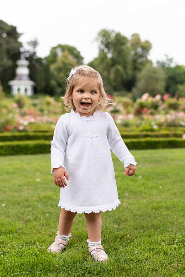 Robe baptême fille blanc cassé en maille tricotée avec bordures faites au crochet à la main. Robe en lainage. Création française Fil de Légende. Photo de la robe portée par une petite fille.