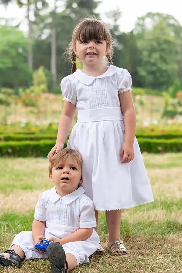 Robe de baptême et de cérémonie pour bébé et petite fille, en piqué de coton blanc et dentelle de coton. Ceinture au choix blanche ou rose. Création française Fil de Légende. Photo de a robe portée par une petite fille avec son petit frère.