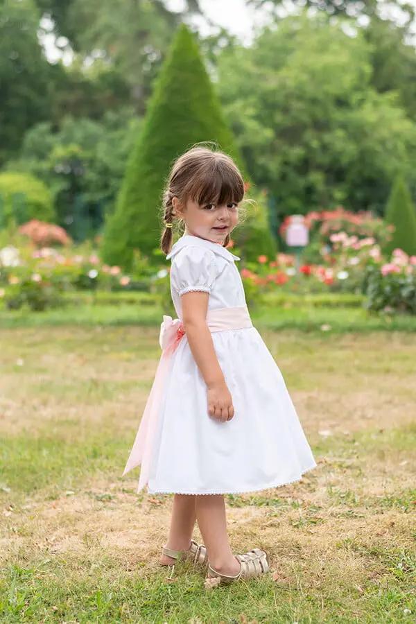Robe de baptême et de cérémonie pour bébé et petite fille, en piqué de coton blanc et dentelle de coton. Ceinture au choix blanche ou rose. Création française Fil de Légende. Photo de robe vue de côté.