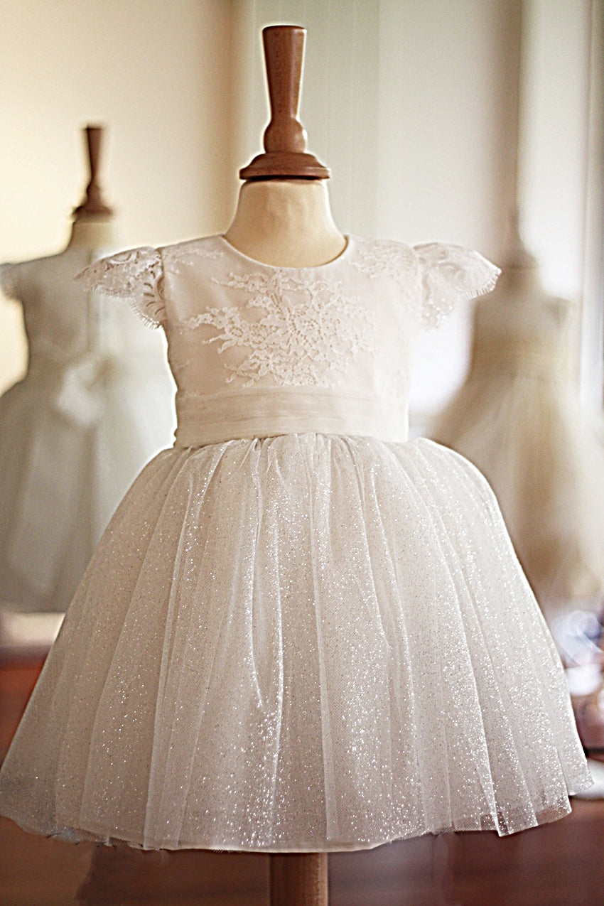 Robe de baptême et de cérémonie pour bébé fille, corsage en dentelle italienne blanc cassé, jupe en tulle pailleté, ceinture en soie. Robe de baptême haut de gamme, création française Fil de Légende, disponible en ligne ou en boutique showroom à Paris – Neuilly‑sur‑Seine.
