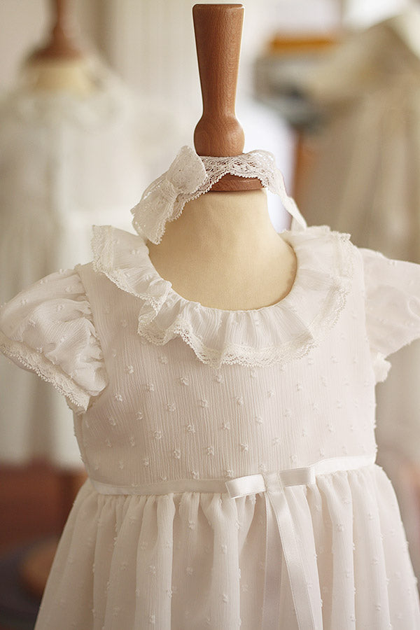 Bandeau cheveux bébé fille en dentelle blanc cassé avec nœud sur le côté. A porter pour un baptême ou une cérémonie. Photo du bandeau porté avec une robe.